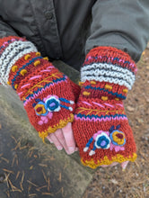 Load image into Gallery viewer, Rust red handknitted wool fingerless mitts with colourful embroidered flowers, worn outdoors. Aster Fingerless Mitts, fair trade, fleece-lined for warmth