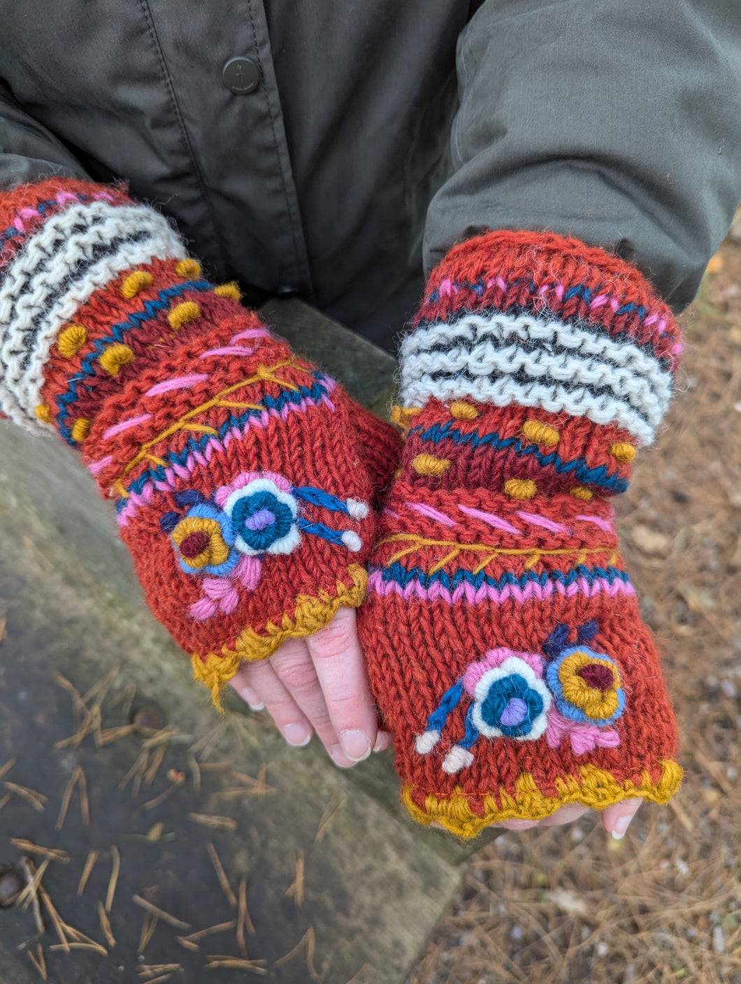 Rust red handknitted wool fingerless mitts with colourful embroidered flowers, worn outdoors. Aster Fingerless Mitts, fair trade, fleece-lined for warmth